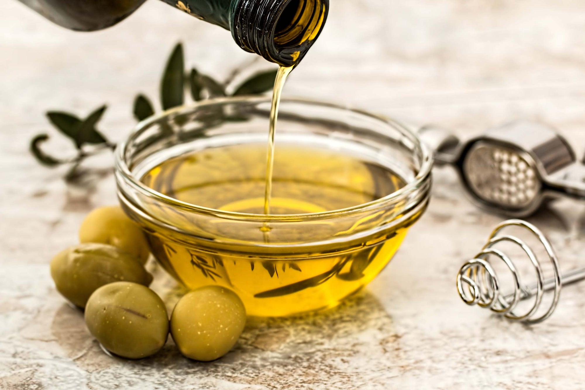 Olive oil being poured from a bottle into a small glass bowl. Olives and other utensils surround the bowl.